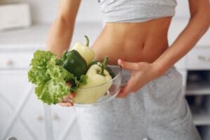 Une femme en tenue de sport tient un bol en verre rempli de légumes verts et de poivrons, debout dans une cuisine lumineuse. Son ventre tonique est visible, ce qui souligne son mode de vie sain et l'importance qu'elle accorde aux aliments qui favorisent la digestion.