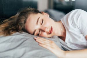 Une jeune femme aux cheveux longs dort paisiblement sur le côté dans son lit, profitant d'un sommeil profond alors qu'elle repose sa tête sur un oreiller gris et porte une chemise blanche. Ses yeux sont fermés et elle a l'air détendue.