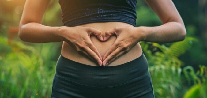 Une personne en tenue de sport se tient à l'extérieur, formant un cœur avec ses mains sur son ventre nu, ce qui suggère l'amour, le soin de la digestion ou la grossesse. La verdure et la lumière du soleil sont visibles en arrière-plan.