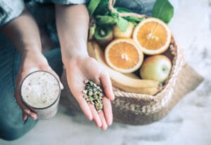 Une personne tient un smoothie et des graines dans ses mains à côté d'un panier tressé rempli de bananes, d'oranges, d'une pomme, d'un kiwi et de légumes verts - des ingrédients parfaits pour favoriser une bonne digestion.