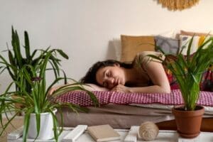 Une femme profite d'un sommeil paisible sur un lit à la literie colorée, entourée de plantes en pot et de coussins moelleux dans une pièce confortable et bien éclairée. Un carnet, de la ficelle et des objets décoratifs sont posés sur une table blanche à proximité.