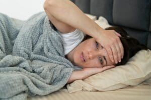 Une femme est allongée dans son lit sous une couverture grise, posant sa tête sur un oreiller et se touchant le front avec une expression douloureuse, indiquant peut-être une nuit troublé ou de la fatigue.