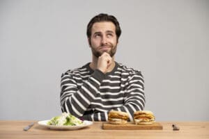 Un homme en chemise rayée est assis à une table, l'air pensif et peut-être un peu affecté par le manque de sommeil. Devant lui, une assiette de salade d'un côté et deux sandwichs de l'autre, comme s'il était en train de décider ce qu'il allait manger.