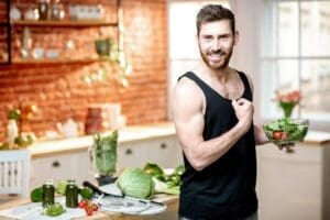 Un homme souriant vêtu d'un débardeur noir montre son biceps fléchi tout en tenant un bol de salade, entouré de légumes frais et de jus vert - une scène parfaite pour ceux qui recherchent de délicieuses recettes végétariennes dans une cuisine lumineuse.