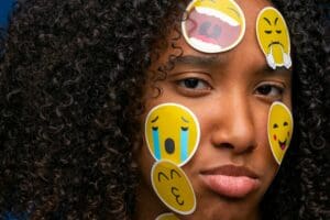 Une personne aux cheveux frisés a l'air triste, avec divers emoji - pleurant, riant, souriant - collés sur son visage, reflétant peut-être les effets du manque d'amour.
