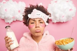 Une femme en sommeil porte un masque de sommeil rose et un sweat à capuche flou, tenant une bouteille de lait et un bol de céréales. Elle arbore des taches roses sous les yeux et une moue boudeuse, avec des nuages duveteux décorant le fond rose.