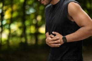 Un homme portant une chemise noire sans manches et une smartwatch se tient le ventre à deux mains, semblant souffrir, peut-être en train de lutter contre les maux de ventre, sur un fond extérieur flou avec de la verdure.