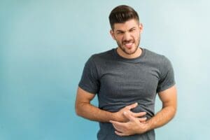 Un homme vêtu d'un t-shirt gris se tient sur un fond bleu clair, serrant son estomac des deux mains et grimaçant de douleur, ce qui suggère un malaise lié à la digestion.